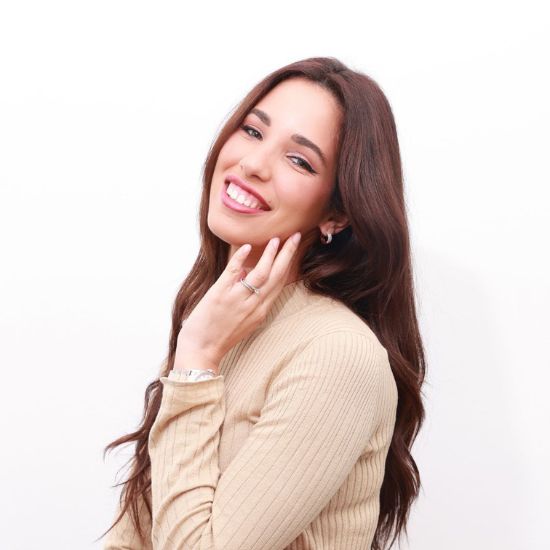 Rocio Montes, a smiling woman with long brown hair, gently touching her face, wearing a beige ribbed sweater against a white background.